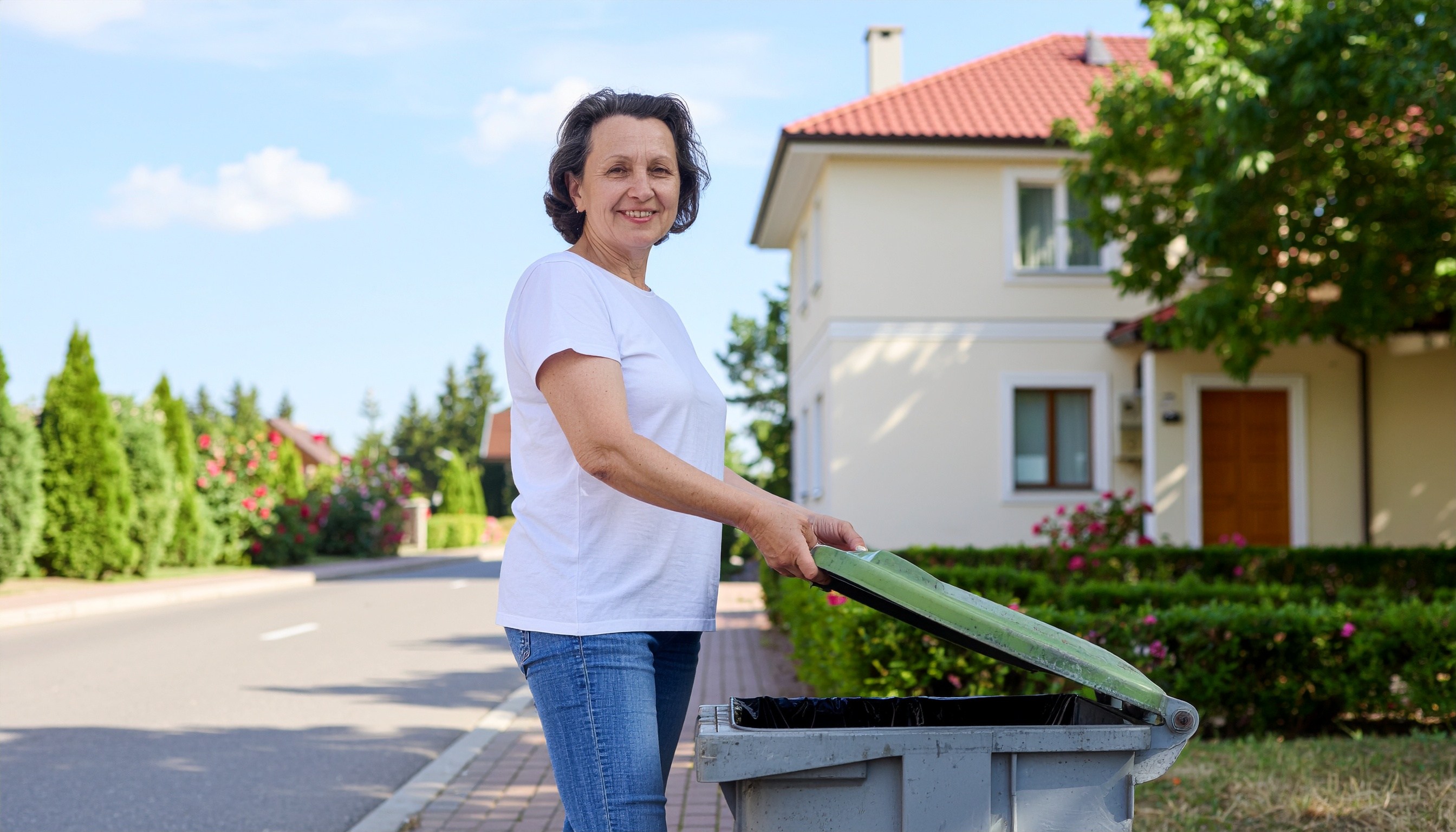 Eine Frau öffnet den Deckel einer an der Straße stehenden Abfalltonne und lächelt in die Kamera.